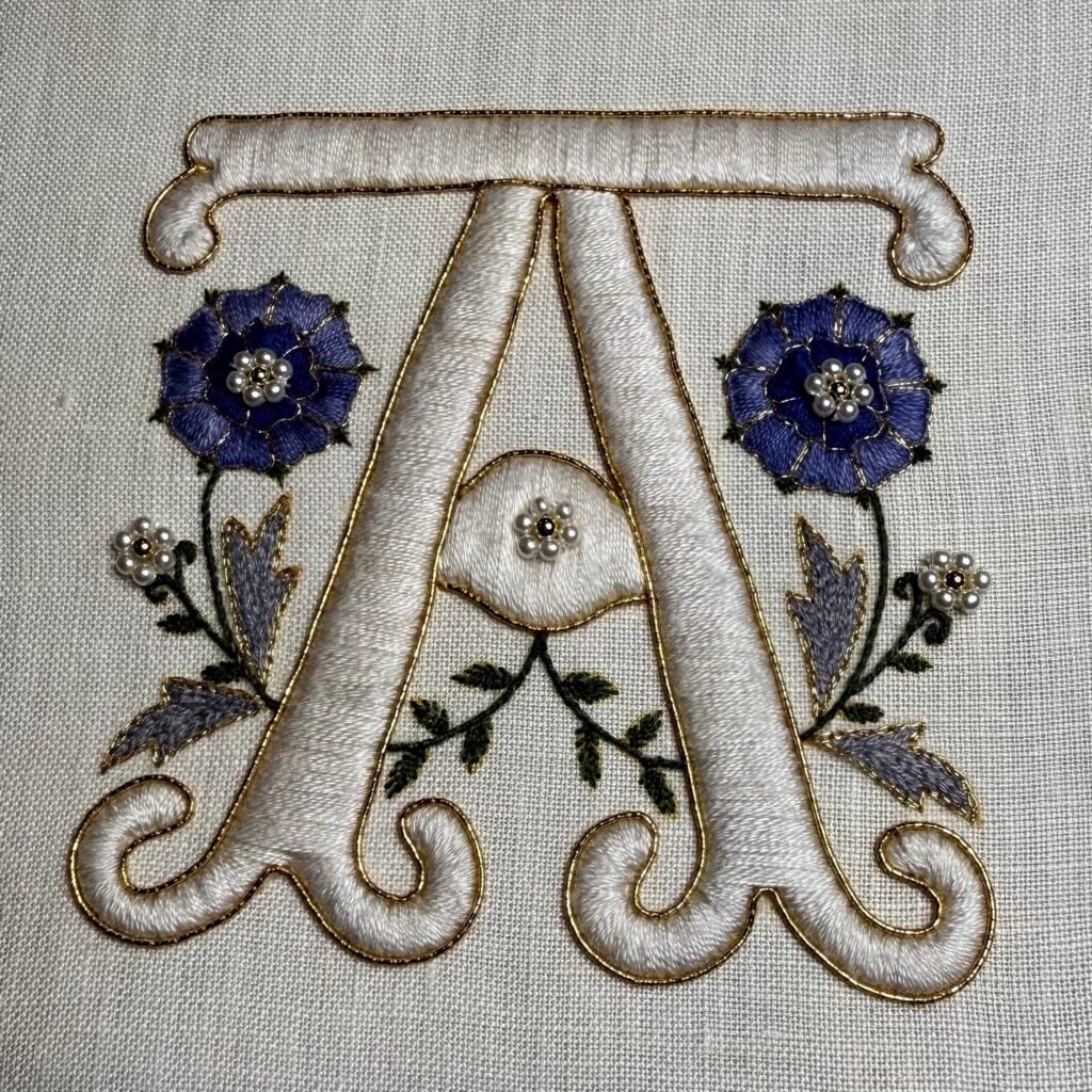A stitched monogram of the letter A in cream satin stitch with blue roses and gold work