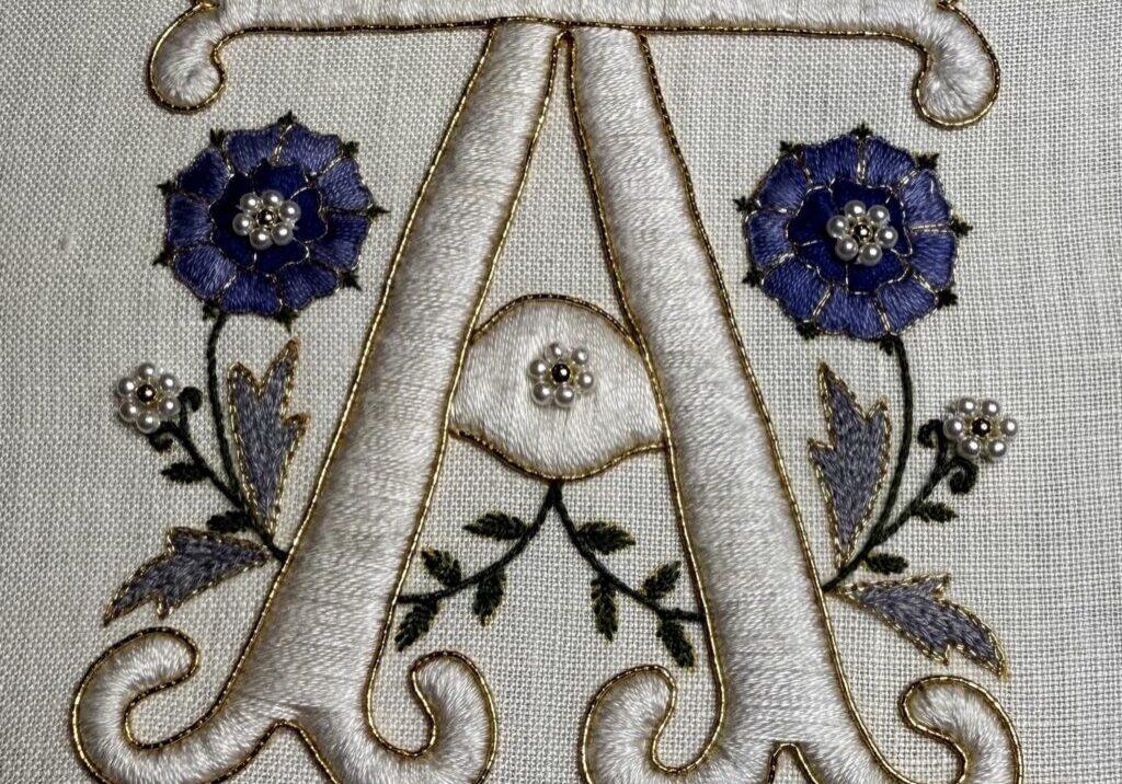 A stitched monogram of the letter A in cream satin stitch with blue roses and gold work